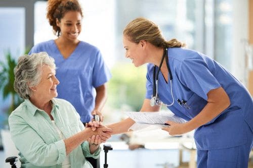 A nurse is leaning over and shaking the hand of a patient in a wheelchair while another nurse stands by ready to push the wheelchair.