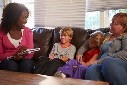 Young female social worker holding pen and pad, sitting on a couch with a mom and two young children.
