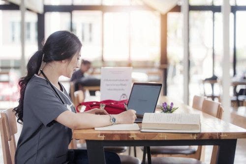Nursing student studying at a table with paperwork and a computer in a well-lit cafe.