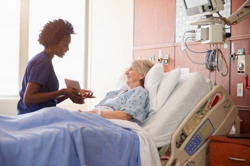 A patient in a hospital bed talks with a nurse holding a tablet.