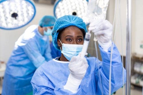 A masked nurse in an operating room prepares an IV.