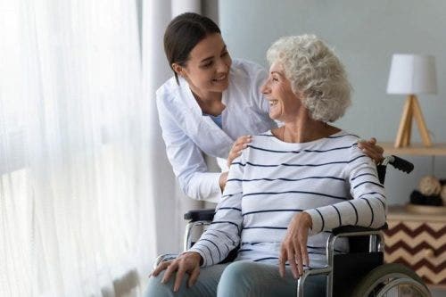 A senior patient sitting in a wheelchair by a window smiles at a nurse while receiving care.