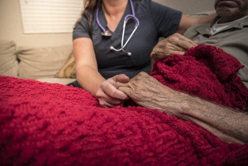 A hospice nurse with a stethoscope around their neck sits at the bedside of a patient and reaches out to hold the patient’s hand.