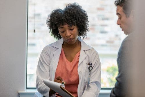 A nurse practitioner gestures at documents on a clipboard while speaking with a patient.
