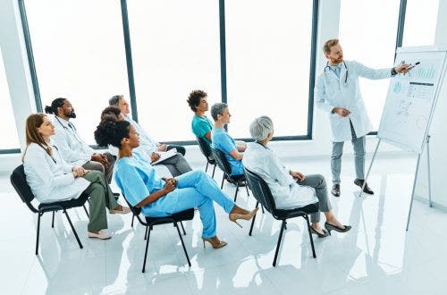A nurse educator lectures at a whiteboard in front of a group of nurses.