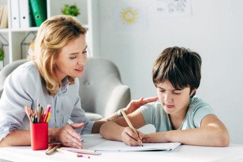 A behavioral therapist sits next to a child drawing with a colored pencil.