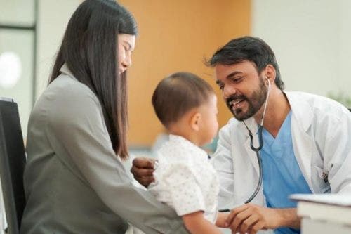 A pediatric nurse uses a stethoscope on a young patient who is sitting on their mother’s lap.