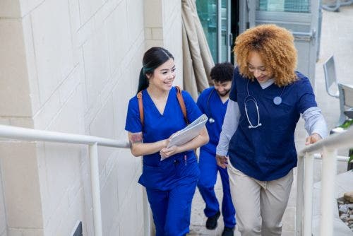 A nurse educator speaks with a smiling nursing student as they climb a stairway in a hospital.