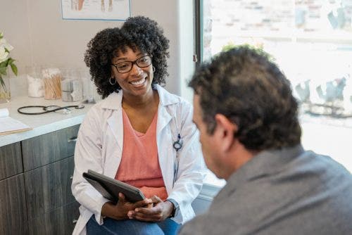 A nurse practitioner holding a tablet smiles while speaking with a patient in an exam room.
