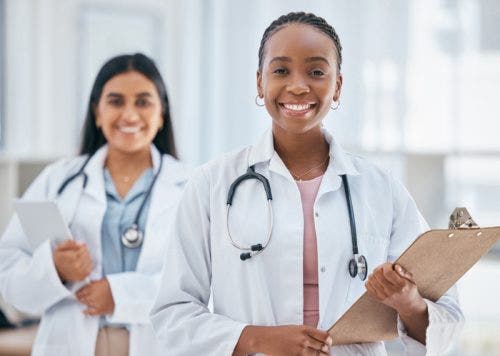 Two nurses wearing white lab coats and holding clip boards, smiling.