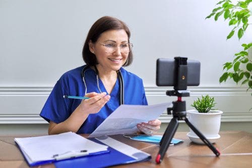 A nurse in blue scrubs talks with a patient on a video call, using a smartphone on a tripod.